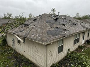 A home with severe storm roof damage showing torn shingles, exposed underlayment, scattered debris, and fallen branches across the roof, highlighting the need for storm damage roof repair.